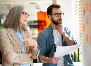 Senior business woman and successful scrum master talking, explaining something using sticky notes, agile methodology for productivity, working together in modern office. Teamwork, scrum concept